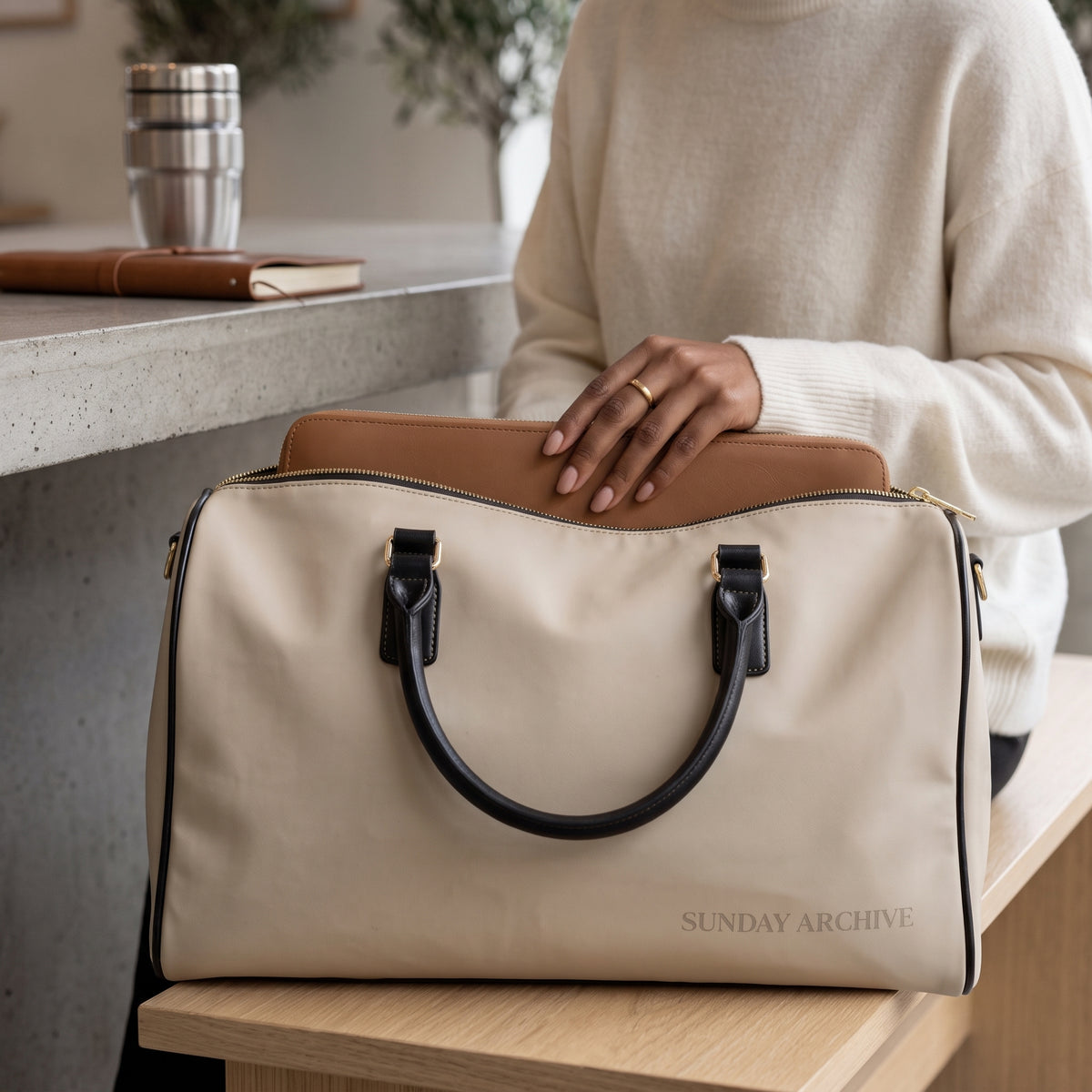 Sunday Archive Off-Duty Weekender in stone with black handles, resting on a cafe counter as a hand tucks a tan leather folio into the open top